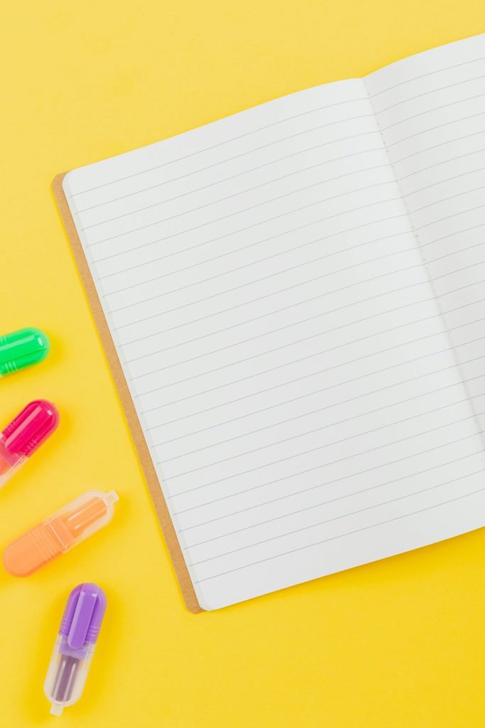 Flat lay of colorful highlighters and an open notebook on a vibrant yellow background.